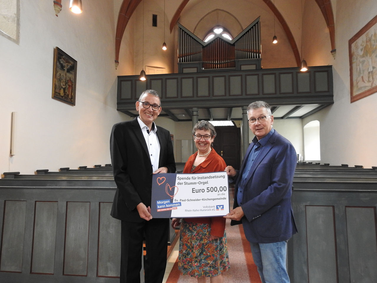 Drei Erwachsene stehen in einer Kirche und halten einen Spendenscheck in der Hand.