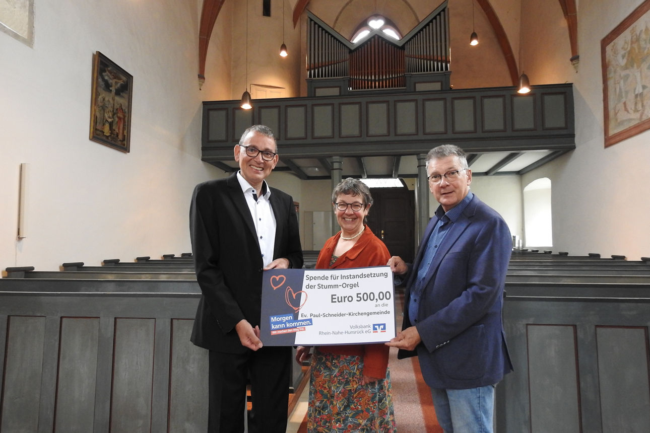 Drei Erwachsene stehen in einer Kirche und halten einen Spendenscheck in der Hand.