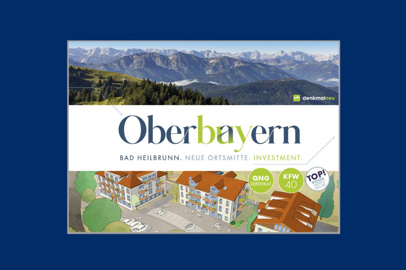 Eine Broschüre mit dem Titel "Oberbayern Bad Heilbrunn. Neue Ortsmitte. Investment." zeigt eine Berglandschaft und ein illustriertes Neubauprojekt. Aufkleber mit "QNG-Zertifikat", "KFW 40" und "TOP! Bester Verkäufer" sind unten rechts angebracht.