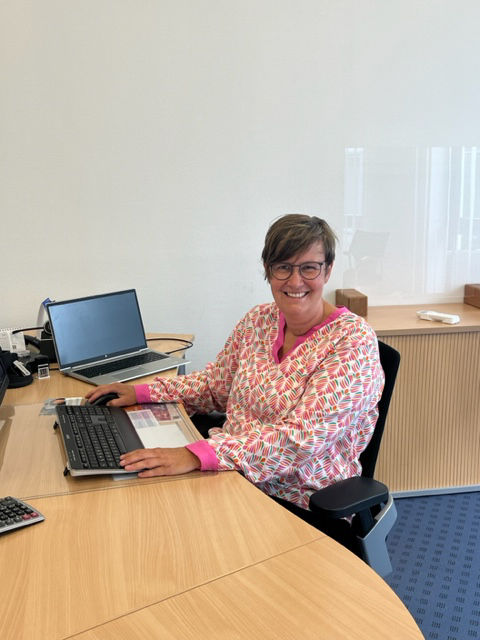 Frau mit Brille sitzt an einer Tastatur an einem Schreibtisch und lächelt