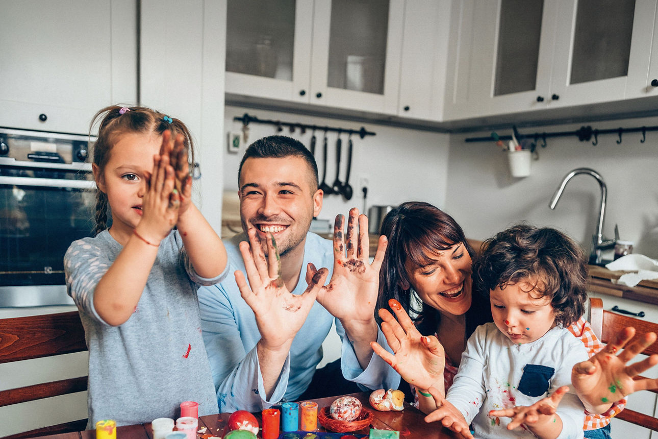 Vierköpfige Familie sitzt am Küchentisch und bemalt gemeinsam bunte Ostereier mit Farben und Pinseln.
