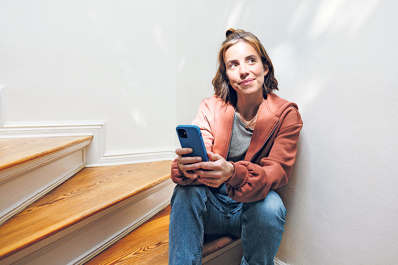 Eine junge Frau sitz mit Handy auf  einer Holztreppe. Die Sonne strahlt ihr ins Gesicht