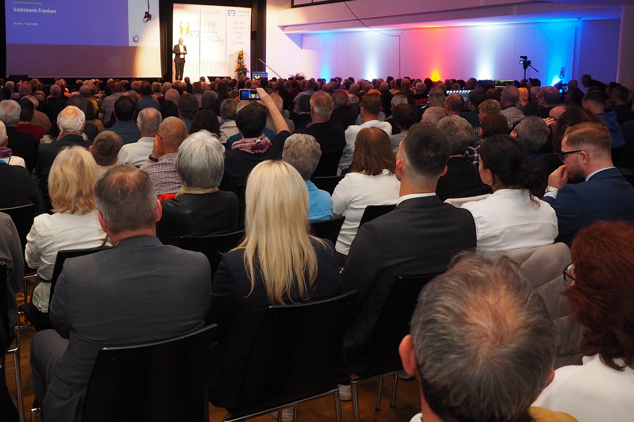 Großes Publikum in Reihen, Blickrichtung zur Bühne mit Leinwand und Vortrag; Saalbeleuchtung mit blauen und orangen Akzenten.