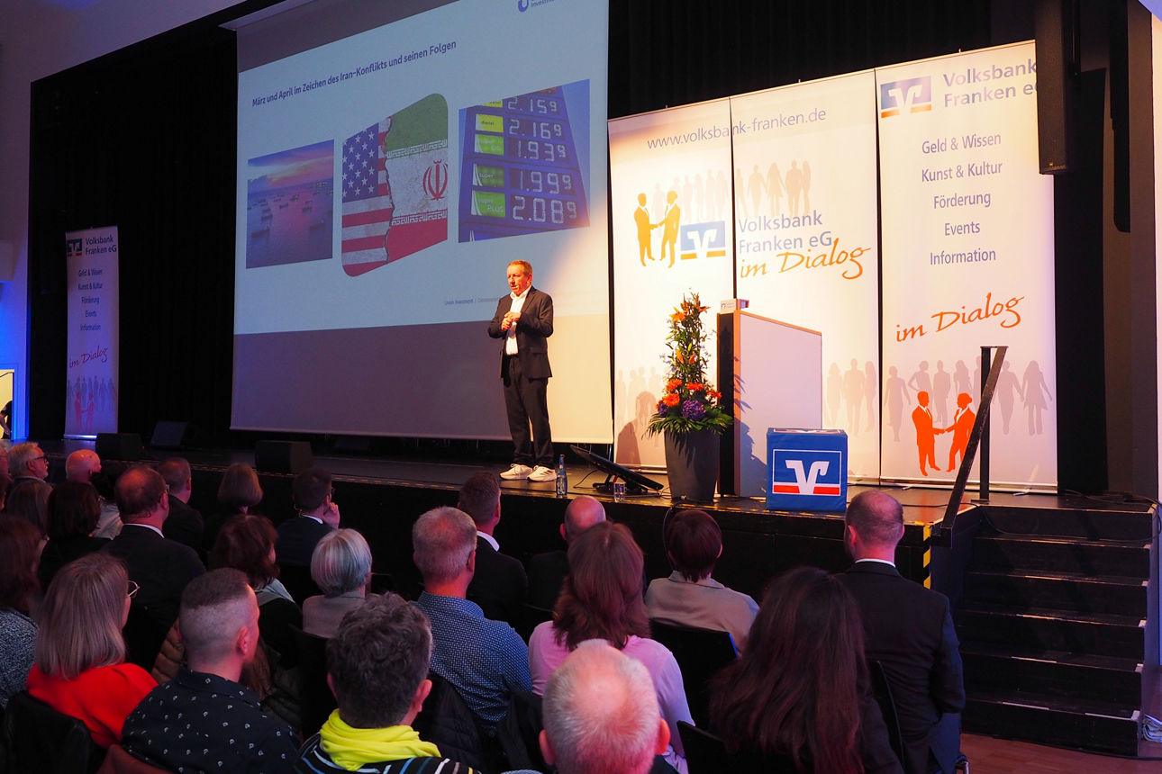 Redner auf einer Bühne vor großer Leinwand mit Grafiken und Flaggenmotiven; rechts Roll‑Ups mit Bank‑Branding, links Publikum