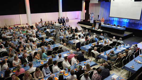Eine große Gruppe von Personen, die in einer geräumigen Halle an Tischen sitzt und sich unterhält und beschäftigt.
