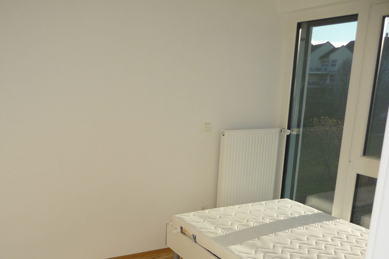 Das Bild zeigt ein beispielhaftes Schlafzimmer im Studentenwohnheim Wellandstraße. Auf der rechten Seite befindet sich eine Fensterfront vor welcher ein Bett platziert ist. 