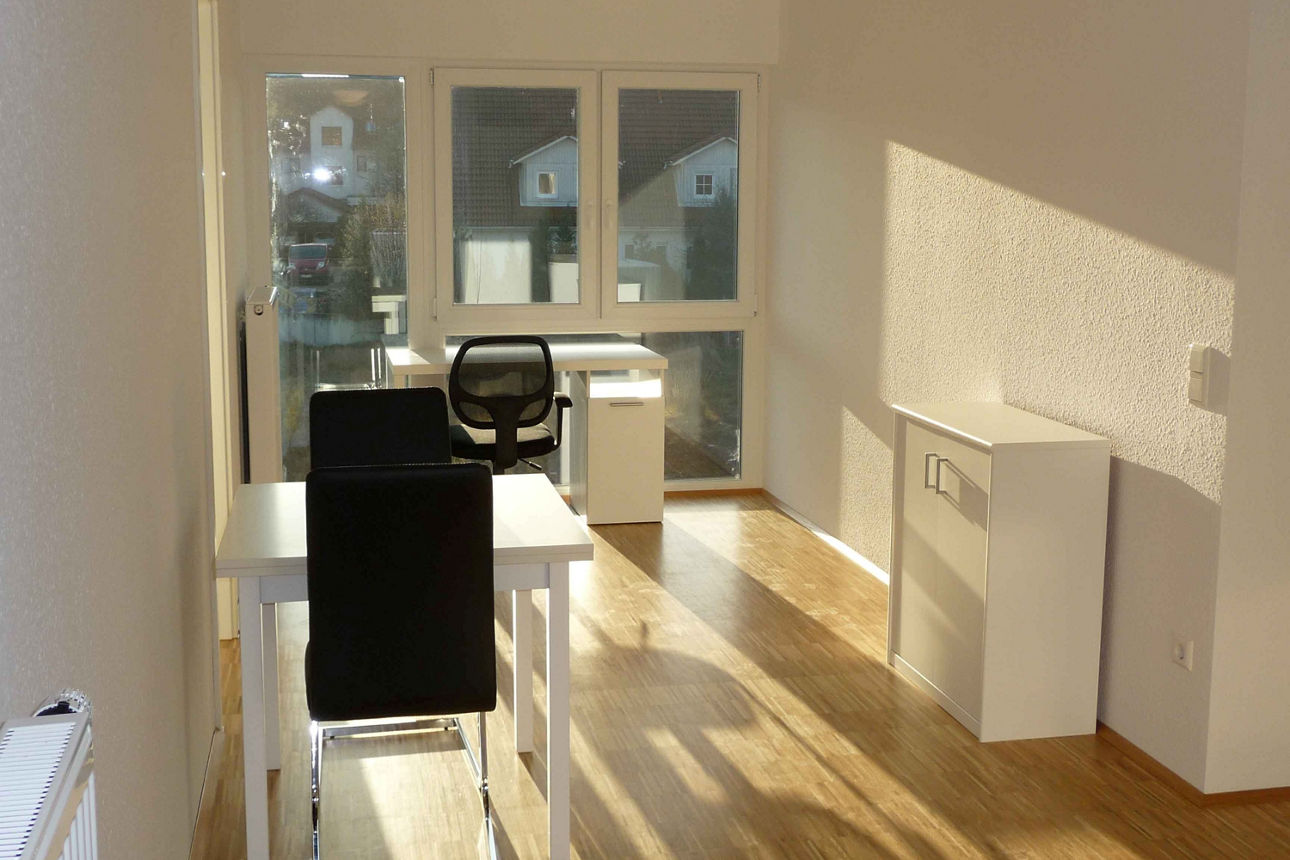 Lichtdurchflutetes Arbeits-/Esszimmer. Direkt vor der großzügigen Fensterfront steht ein weißer Schreibtisch mit einem schwarzen Stuhl. Etwas weiter hinten befindet sich ein kleiner weißer Esstisch mit zwei schwarzen Stühlen. An der langen Wand rechts steht ein kleines, weißes Sideboard.  