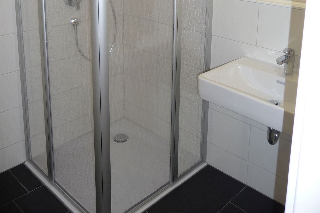 Das Bild zeigt ein beispielhaftes Badezimmer im Studentenwohnheim Wellandstraße. Links befindet sich die Dusche. Rechts daneben ein Waschbecken und darüber ein Spiegel mit Beleuchtung. 