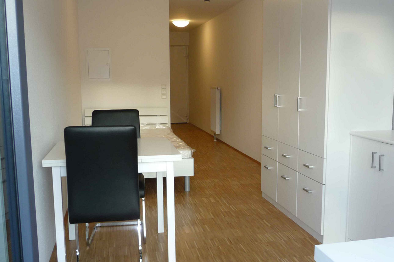 Blick von der Fensterfront zur Tür. Auf der rechten Seite befindet sich der weiße Schreibtisch mit schwarzem Schreibtischstuhl. Weiter hinten links befindet sich die kleine Essecke mit weißem Esstisch und zwei schwarzen Stühlen. Auf der rechten Seite reihen sich ein Sideboard und ein hoher Schrank aneinanander. 