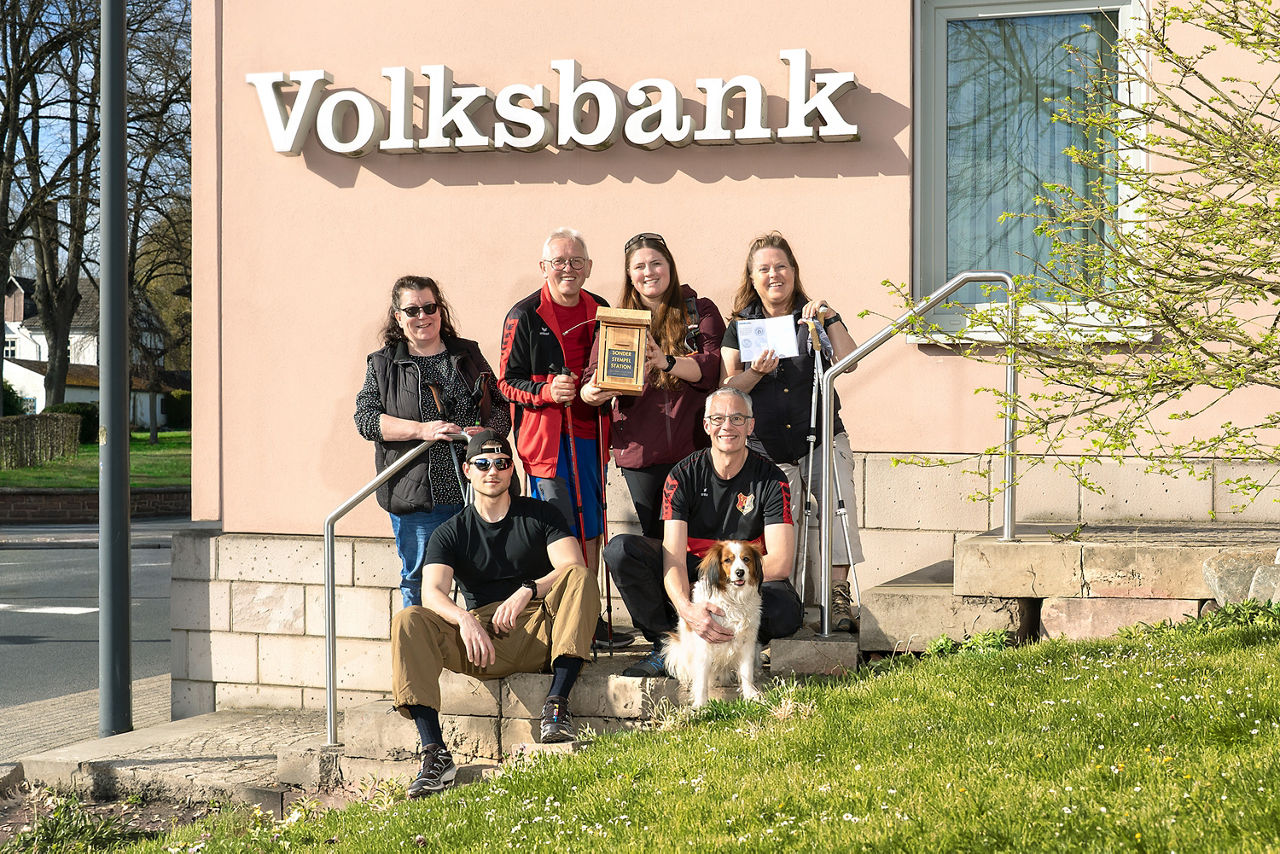 Eine Gruppe von sechs Personen und ein Hund posieren lächelnd vor einem Volksbank-Gebäude in der Sonne.