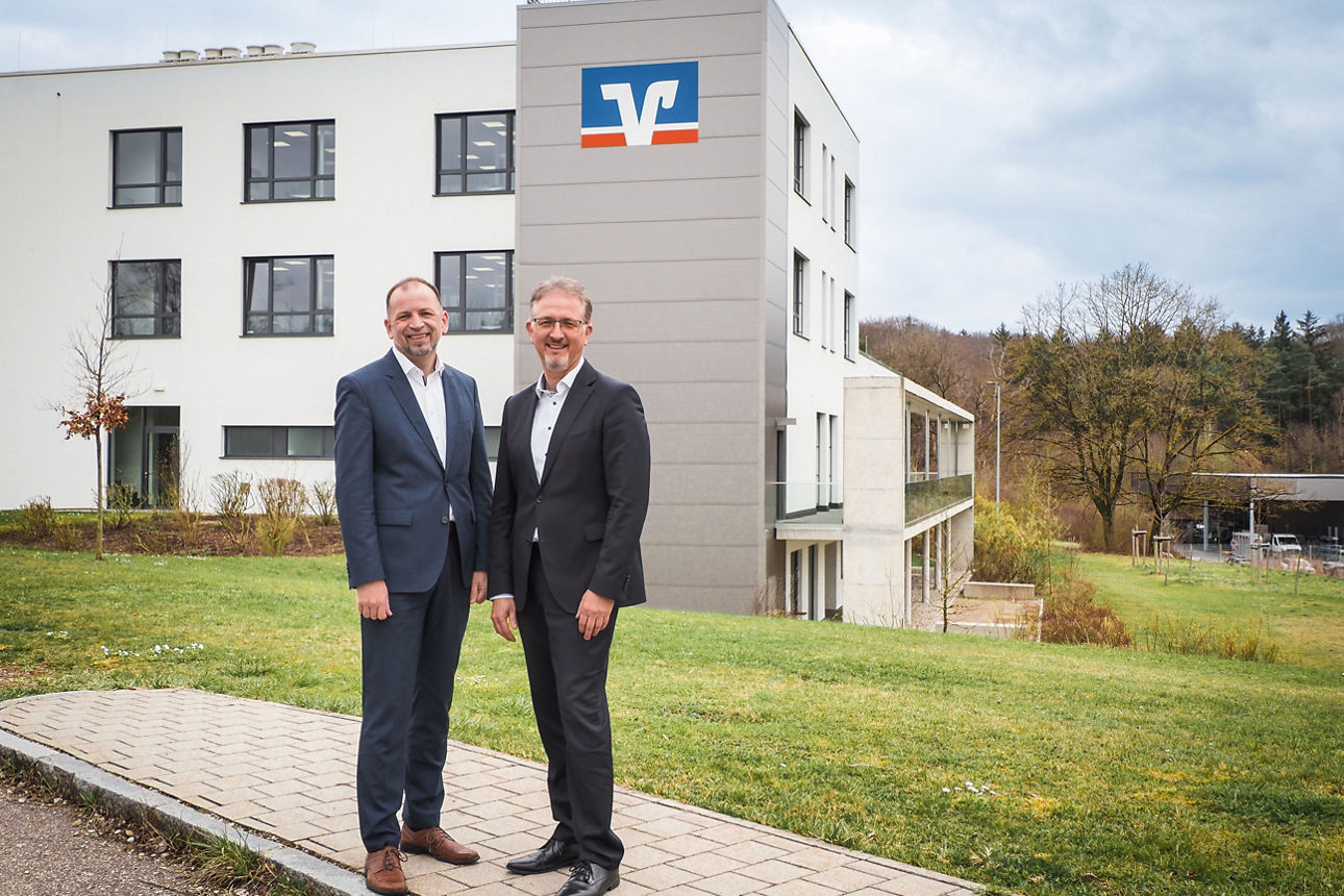 Vorstand Bernd Bengel und Roland Gieß stehen vor einem Verwaltungsgebäude mit VR-Logo im Hintergrund