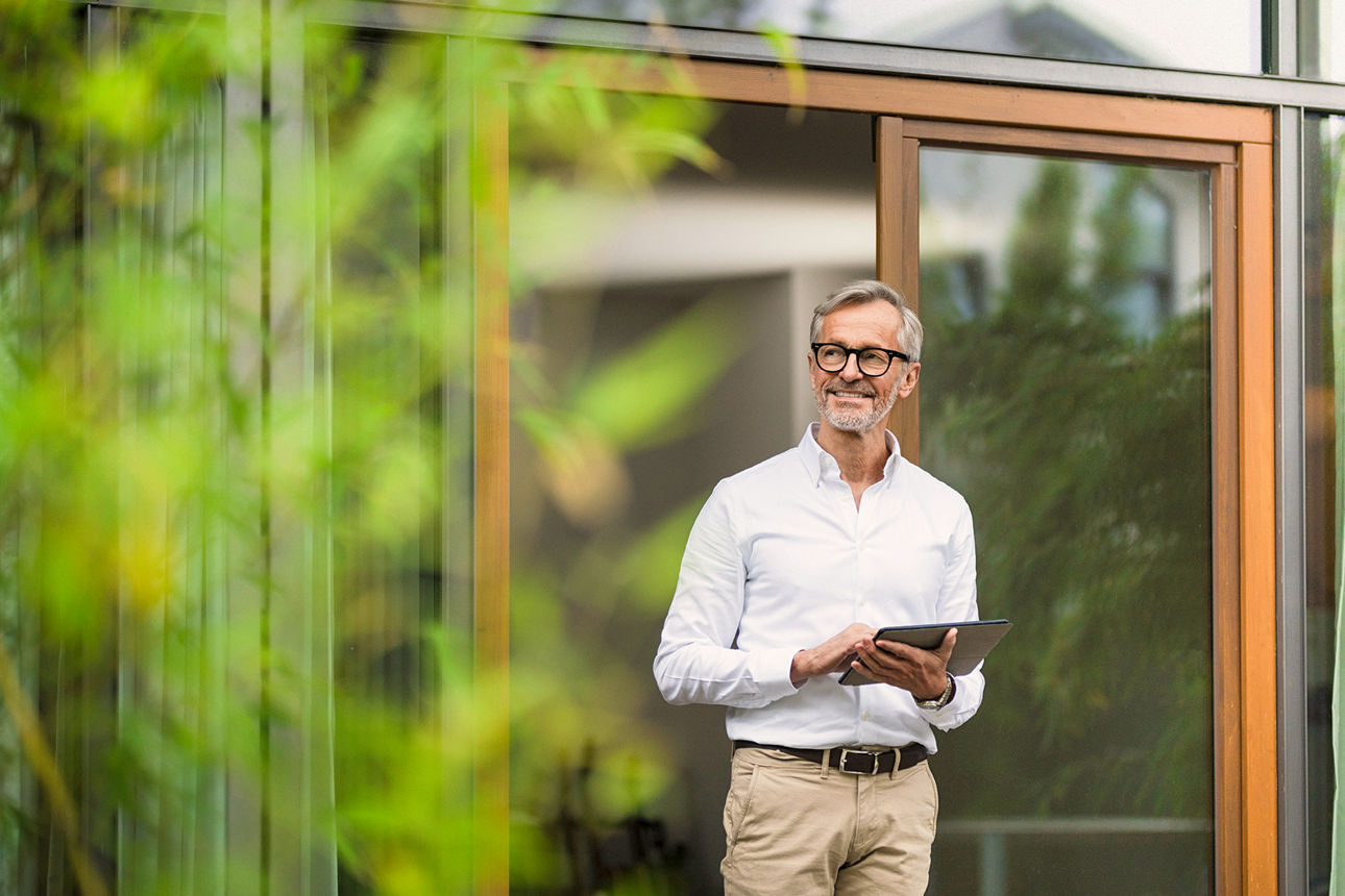 Lächelnder älterer Mann mit grauem Haar, der ein Tablet vor seinem modernen Designhaus im Bambusgarten hält, Deutschland