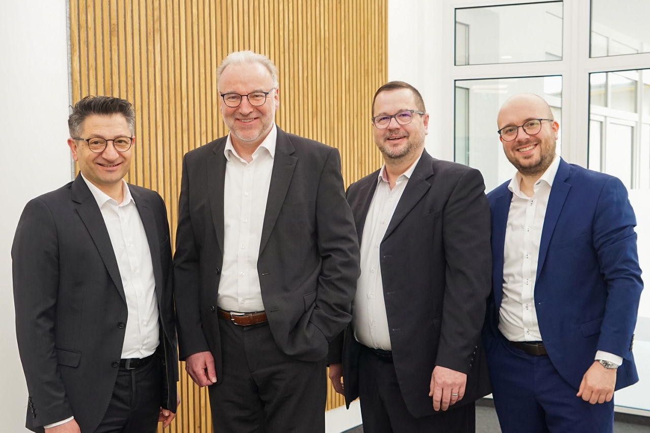 Vier Männer mittleren Alters stehen vor einer modernen Wand in Holzoptik. Sie tragen alle einen Anzug