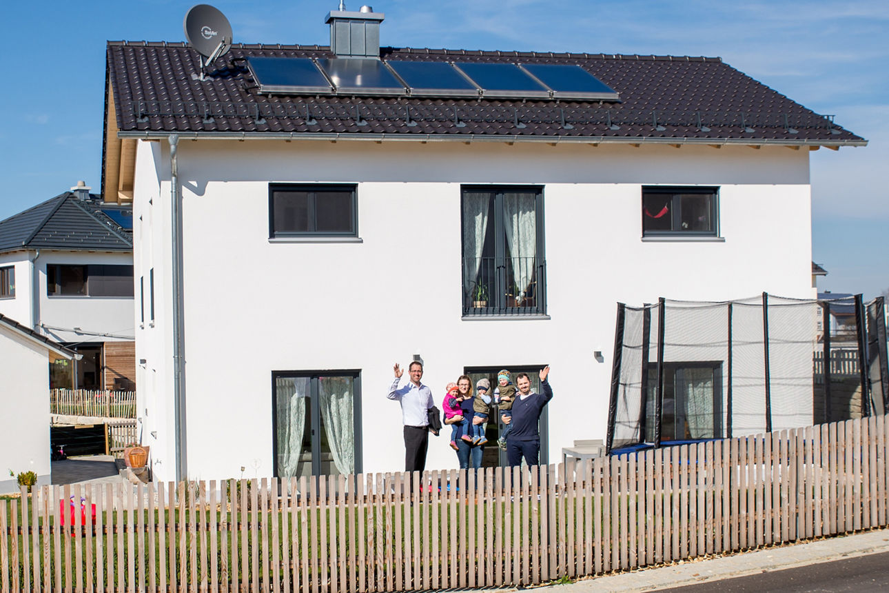 Eine Familie mit drei kleinen Kindern, Mutter und Vater steht mit ihrem Bankberater vor einem neu gebauten weißen Haus. Im Vordergrund der Garten mit einem Holzzaun und rechts ein Trampolin
