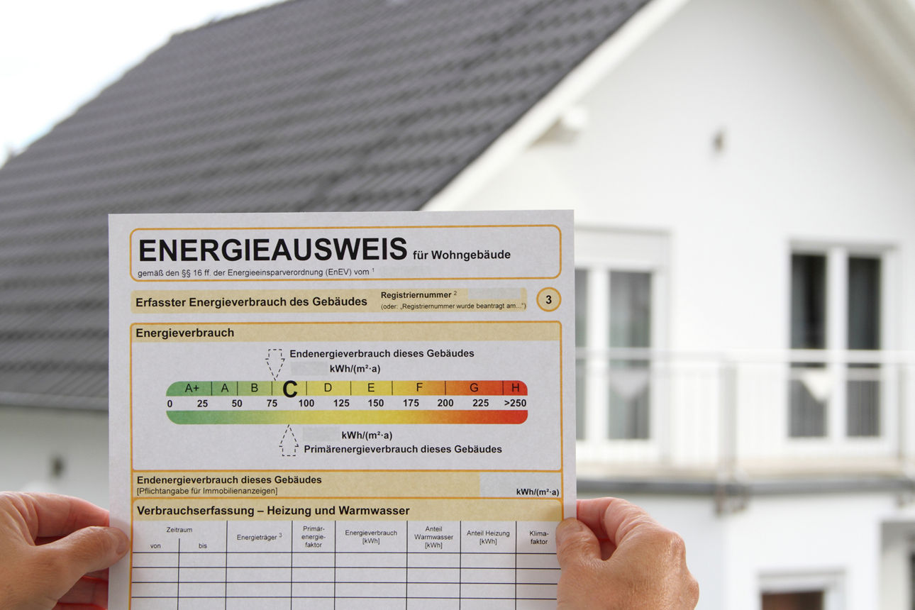 Hände halten Energieausweis in die Höhe, im Hintergrund ein weißes Haus_Getty1433543192