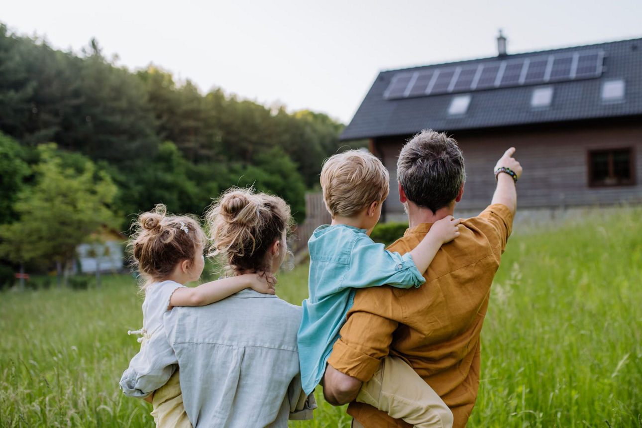 Bild einer jungen Familie, die über eine grüne Wiese geht und auf ihr Haus zuläuft