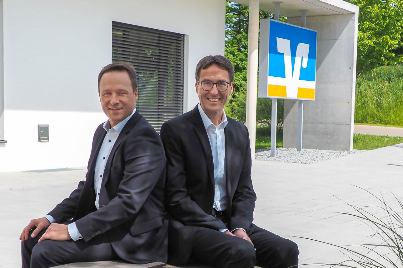 Zwei Männer sitzen lächelnd neben dem VR Bank Logo