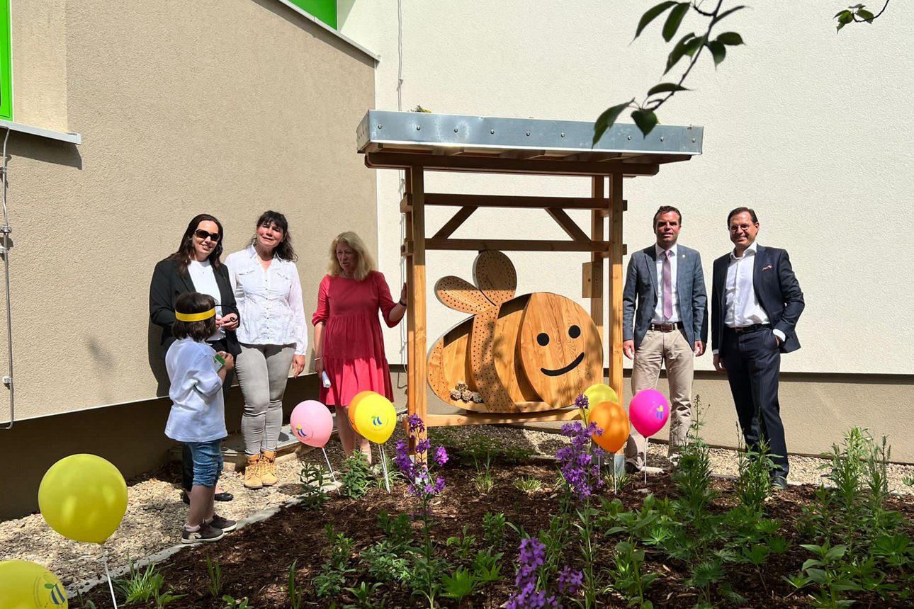 Fünf Erwachsene und ein Kind stehen neben einer großen Holz-Wildbienennisthilfe, umgeben von Blumen und bunten Luftballons.