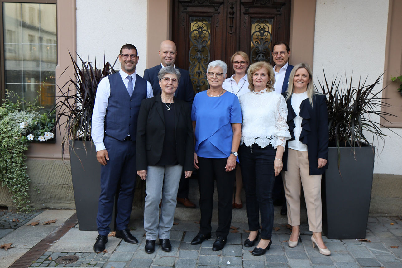 Vor dem Eingang eines Gebäudes stehen acht Personen die in die Kamera lächeln 