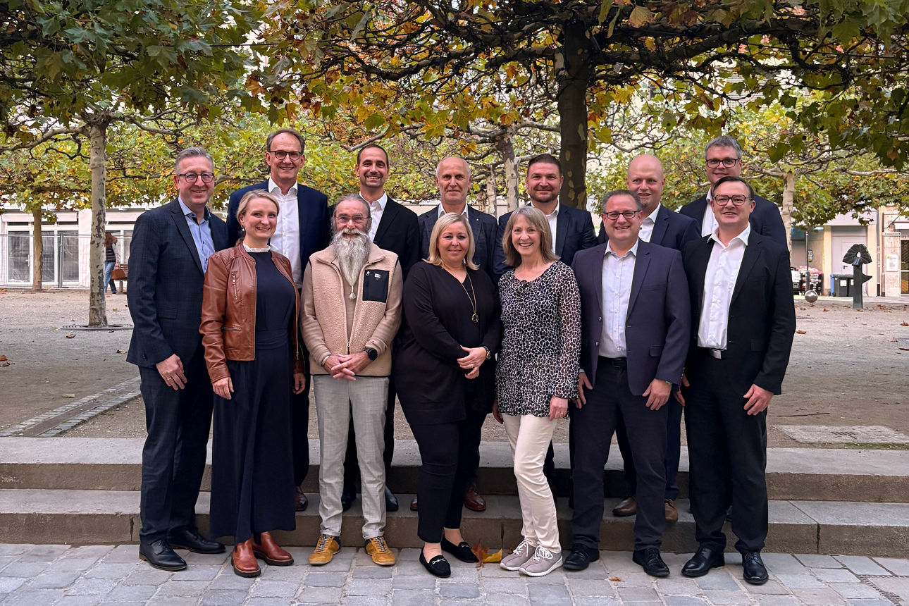 Vor mehreren Bäumen auf einem gepflasterten Platz  steht eine Gruppe von Personen die festlich gekleidet ist