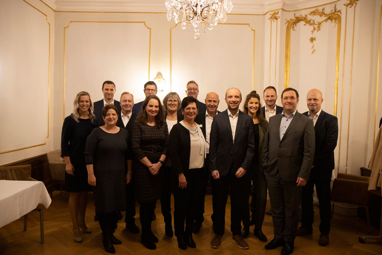 In einem altehrwürdigen Saal stehen einige Personen und werden von einem Kronleuchter beschienen