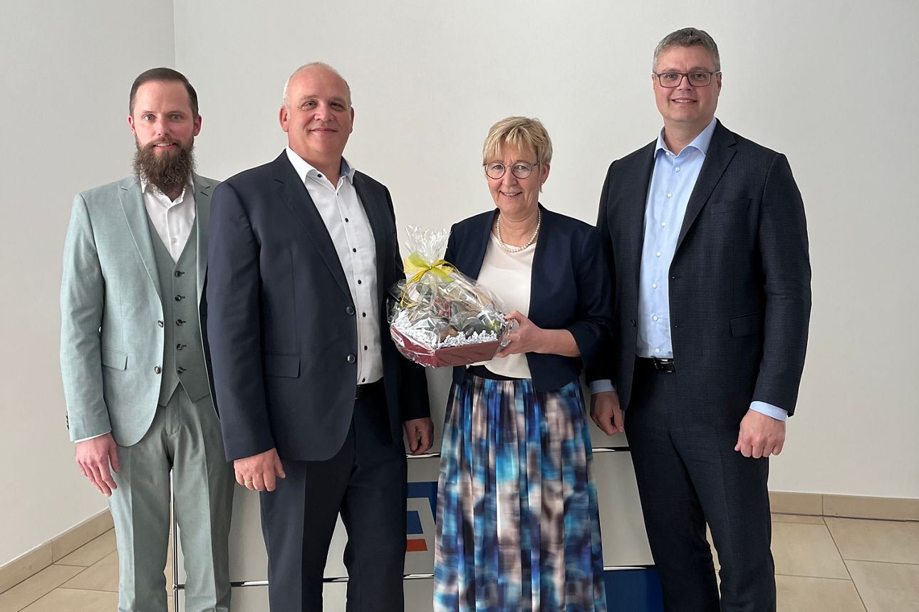 Drei Männer und eine Frau stehen vor einer weißen Wand, die Frau hält einen Präsentkorb in der Hand 