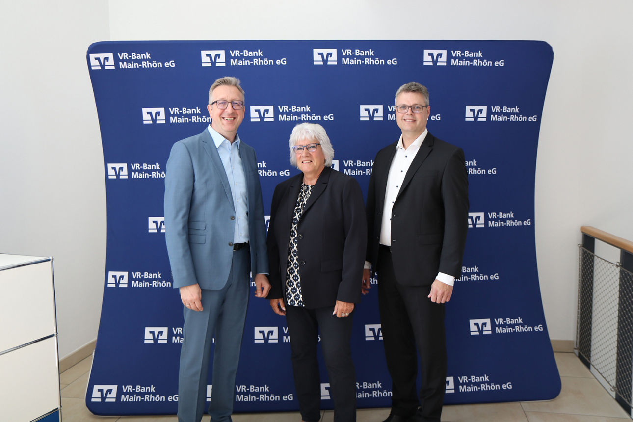 Vor einer blauen Wand mit vielen VR-Bank Maiin-Rhön Logis stehen drei Personen, die in die Kamera lächeln 