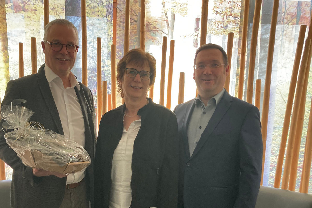 Vor einer Sitzecke steht eine Frau und zwei Männer, der Mann zur linken einen Präsentkorb in der Hand 