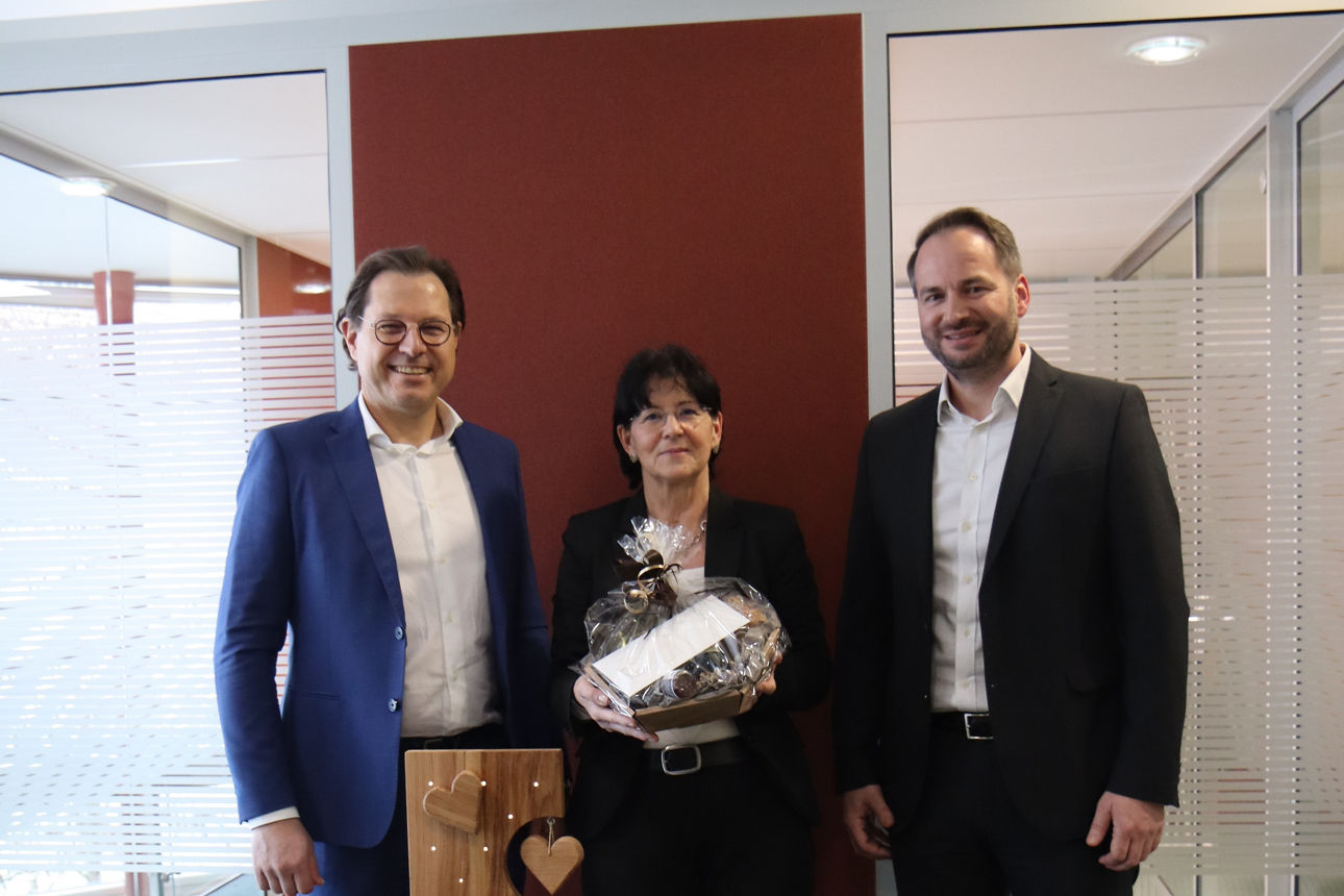 Vor einer roten Wand stehen zwei Männer und eine Frau, die einen Präsentkorb in der Hand hält 