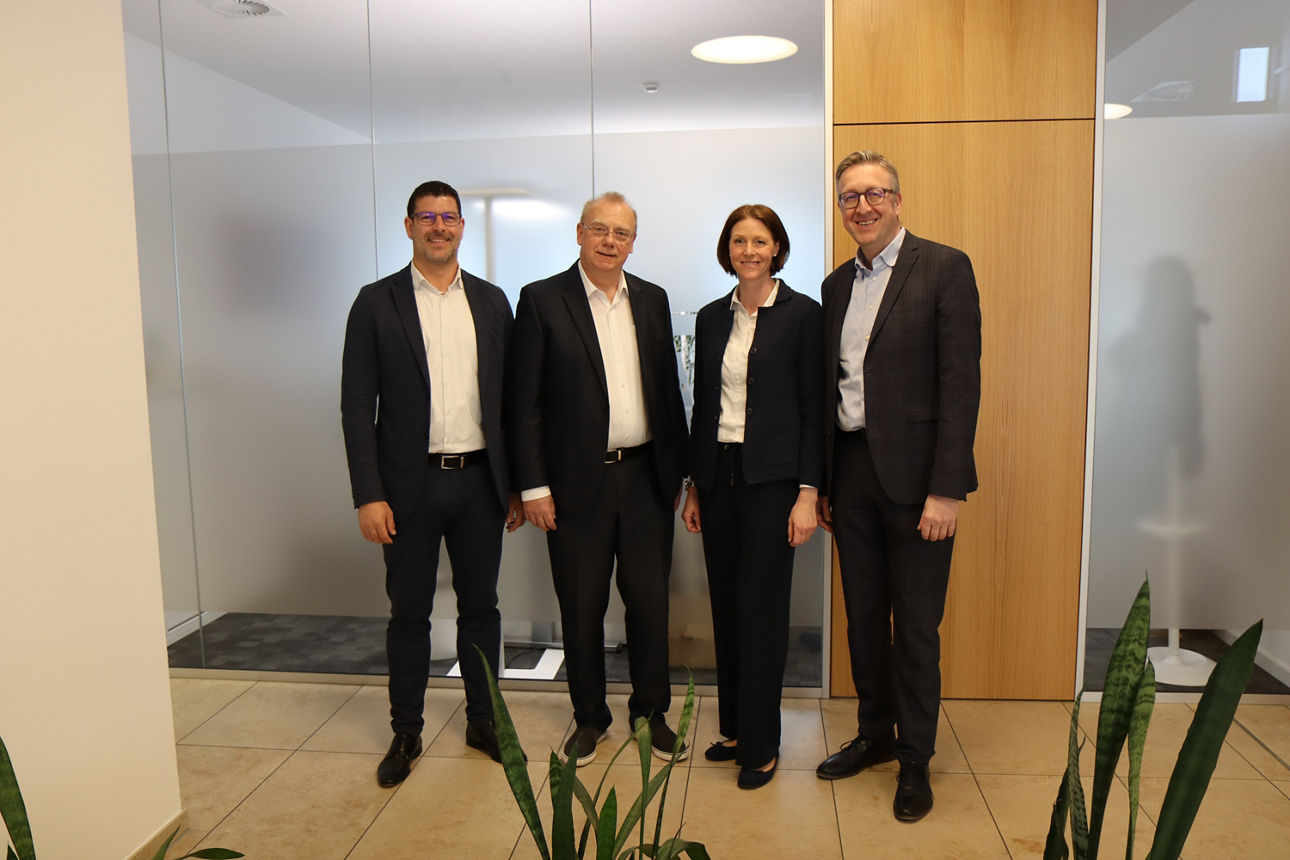 Vor einer Glaswand stehen drei Männer und eine Frau in Anzug 