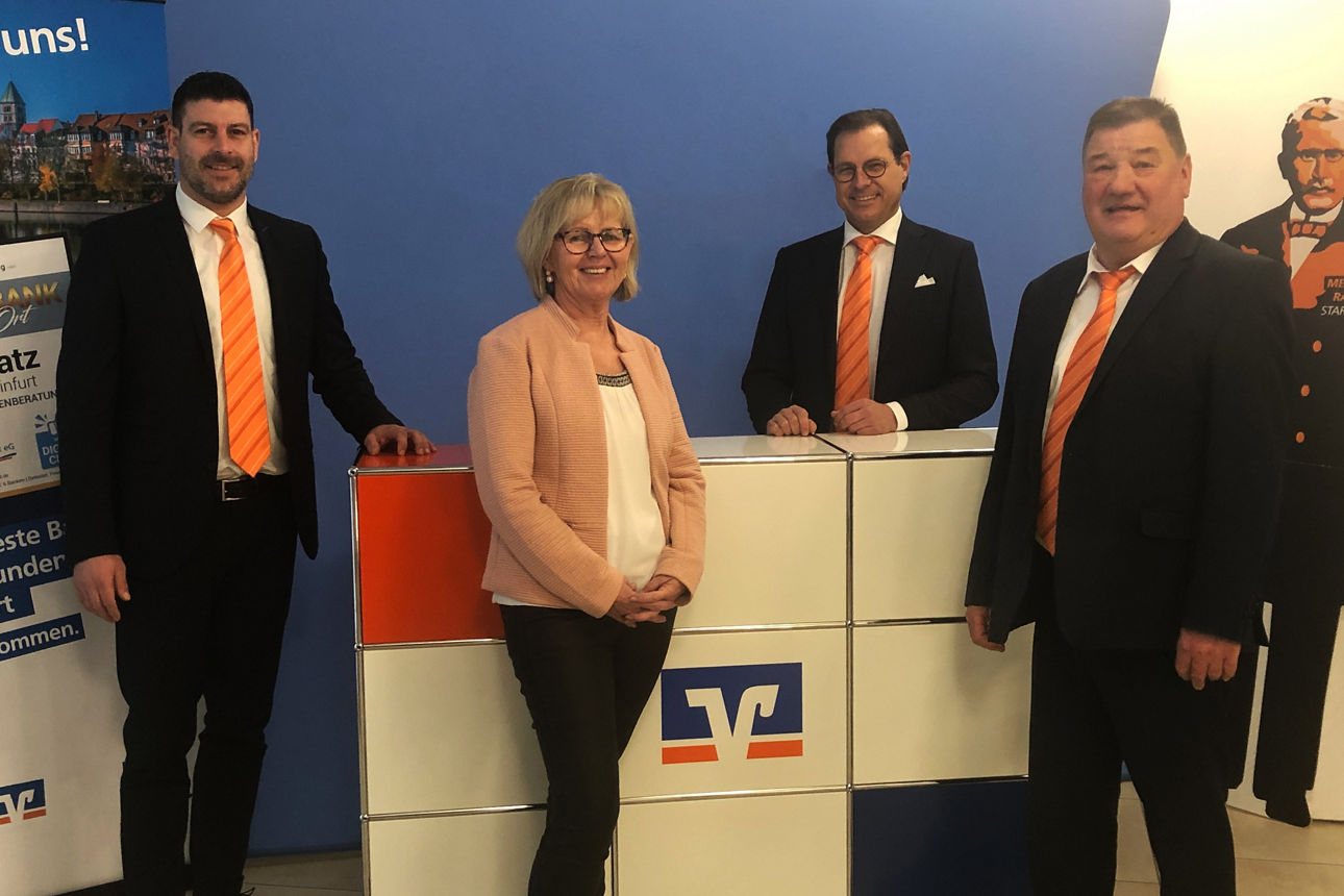 Drei Männer und eine Frau stehen vor einer blauen Wand und einem Pult mit VR-Bank Logo