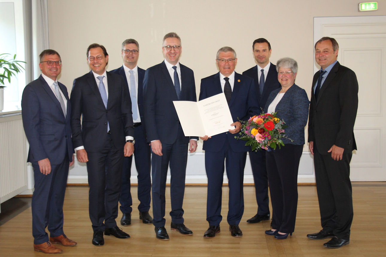 Acht Personen stehen in einem Raum; ein Mann in der Mitte hält eine Urkunde, eine Frau daneben einen Blumenstrauß