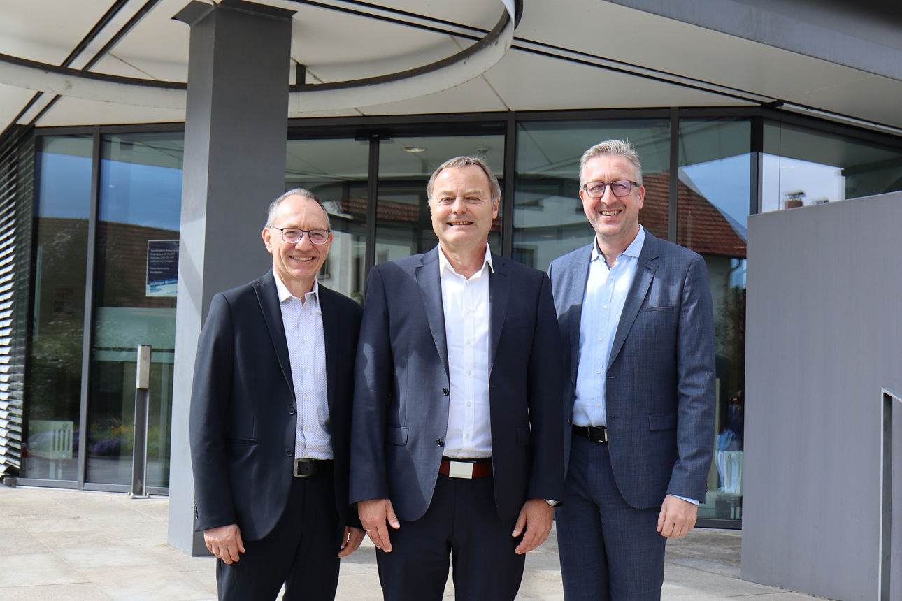 Drei Männer in Anzügen stehen vor einem modernen Gebäude mit einer runden Überdachung und lächeln in die Kamera.