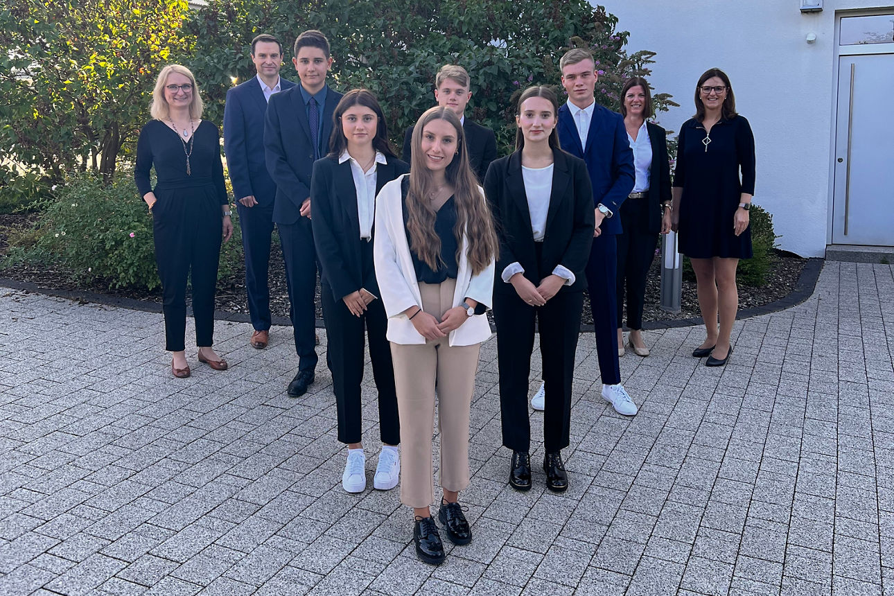 Vor einer Hauswand und einem Busch stehen schechs junge Erwachsene und Jugendliche, sowie vier Erwachsene an der Seite 