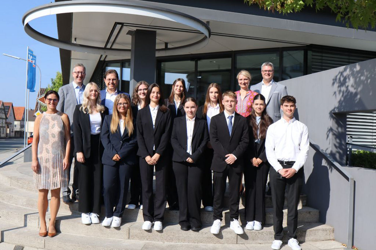 Zehn Auszubildende stehen mit einigen Führungskräften auf der Treppe eines Bankgebäudes 