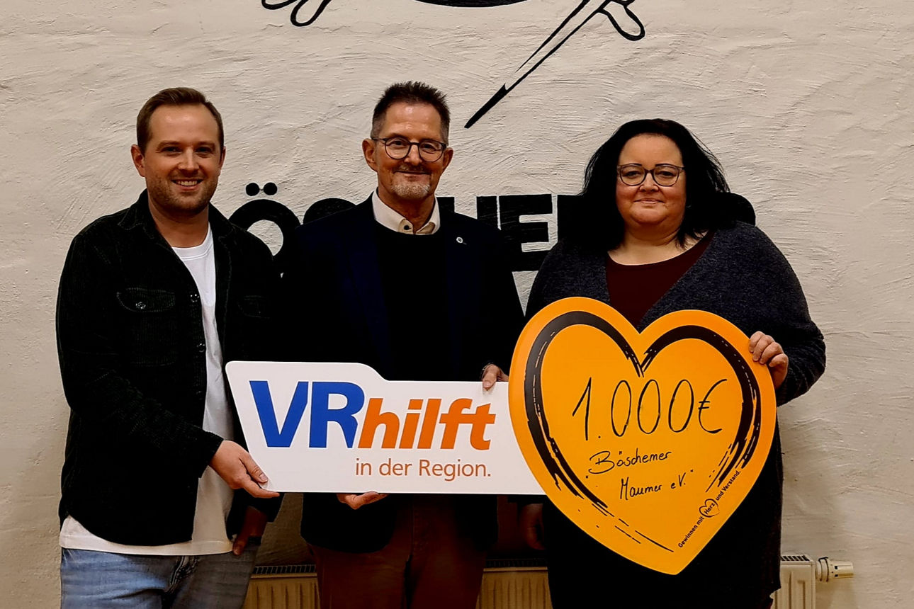 Vor einer Wand stehen drei Personen, die ein Schild mit "VRhilft" und 1.000 Euro "Böschemer Maumer e.V." in der Hand halten