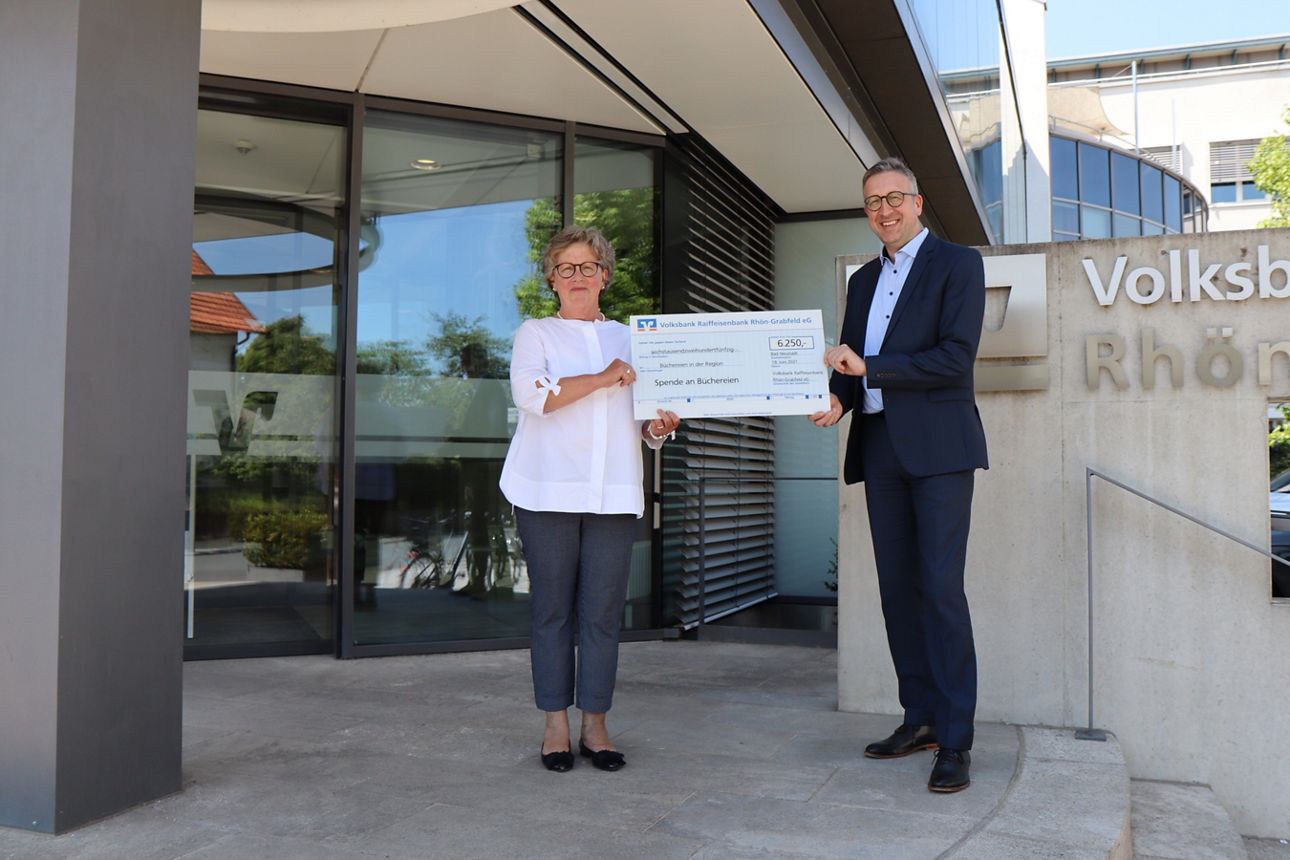 Ein Mann und eine Frau stehen vor einem Bankgebäude und halten einen Spendenscheck in der Hand 