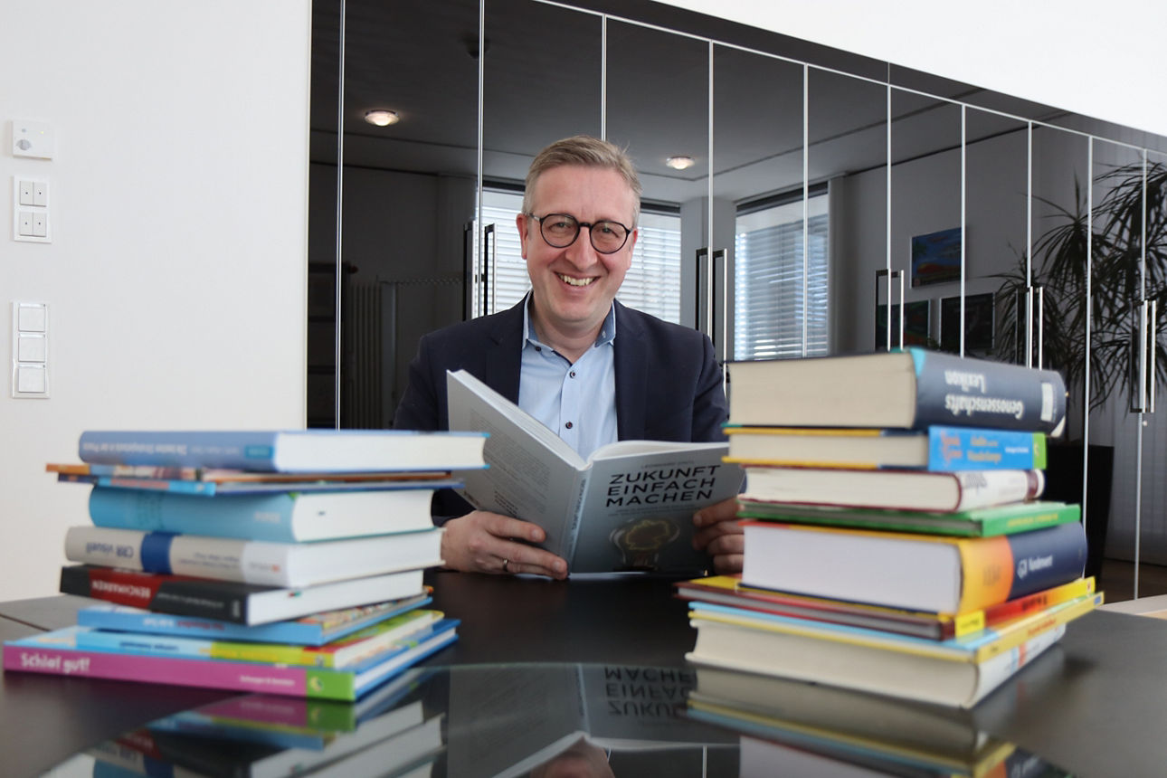 Ein Mann sitzt vor einem Tisch auf dem viele Bücher liegen und hält ein offenes Buch in der Hand