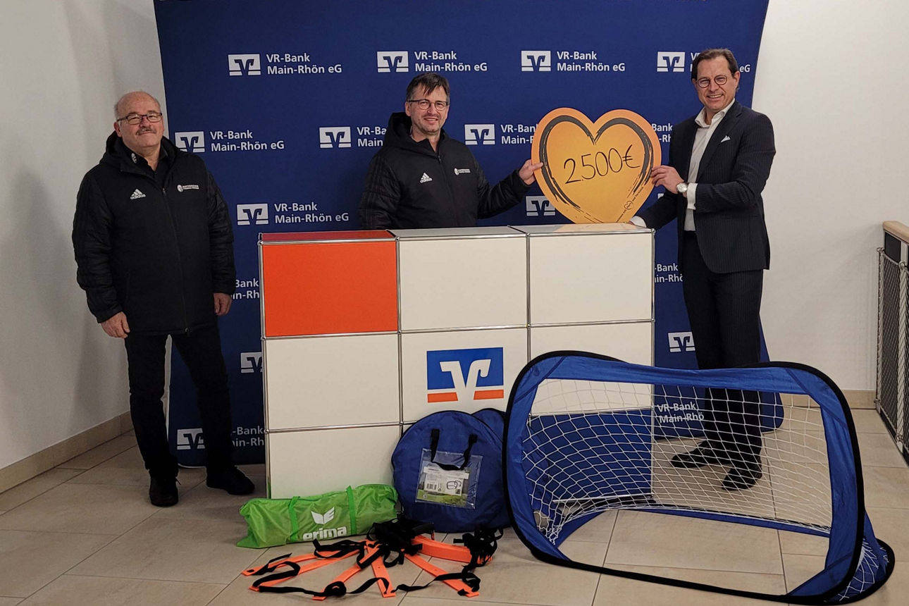 Drei Männer stehen vor einer Wand, zwei halten ein großes Herz-Symbol und vor ihnen liegen Sportartikel