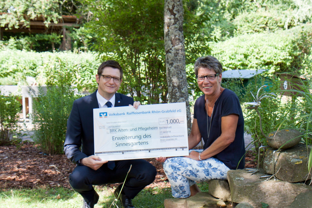 Ein Mann und eine Frau halten in einem Garten einen Spendenscheck in der Hand 