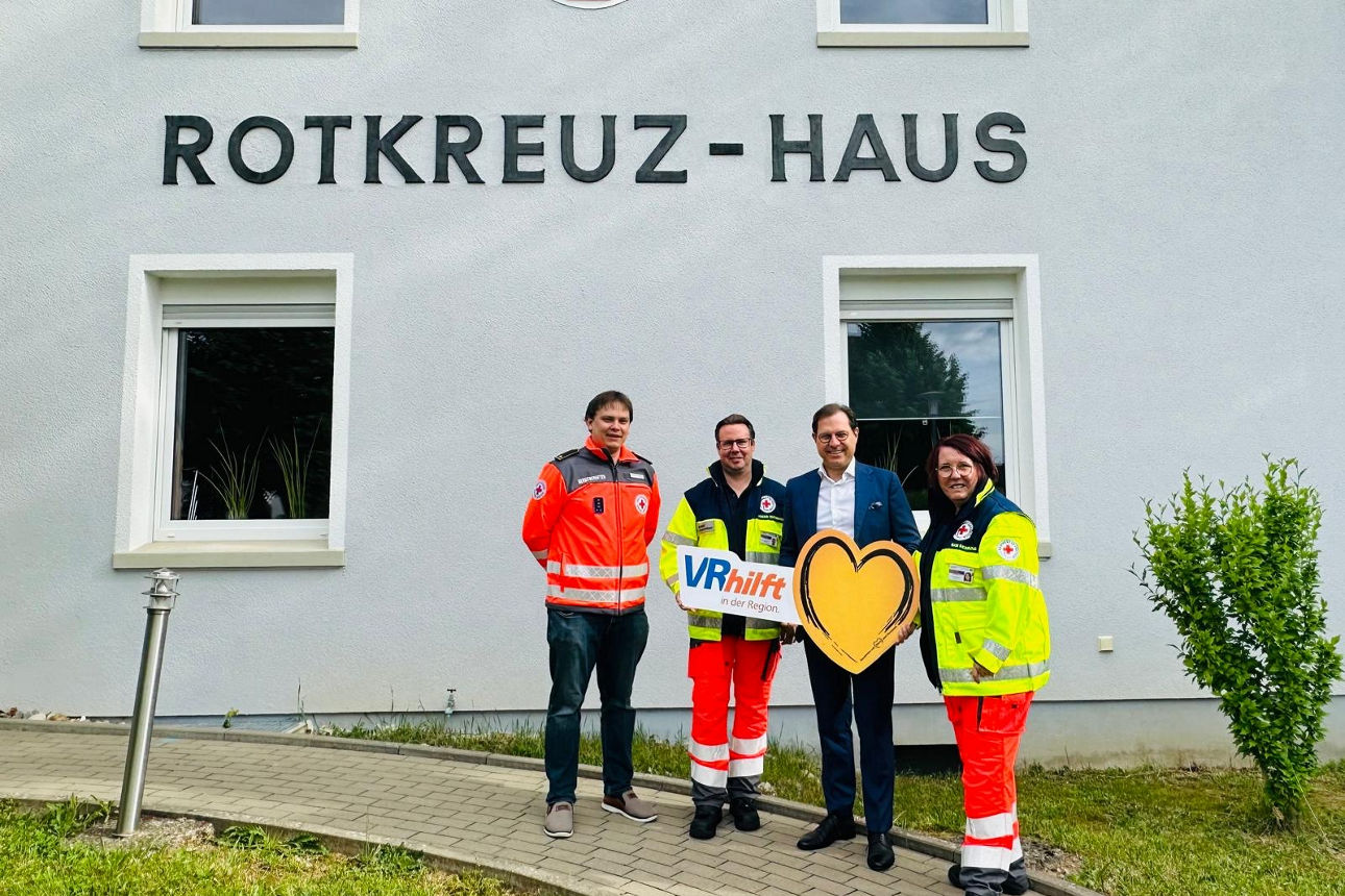 Vier Personen stehen vor einem Gebäude mit der Aufschrift „Rotkreuz-Haus“, zwei in Rettungsdienstkleidung halten ein Schild