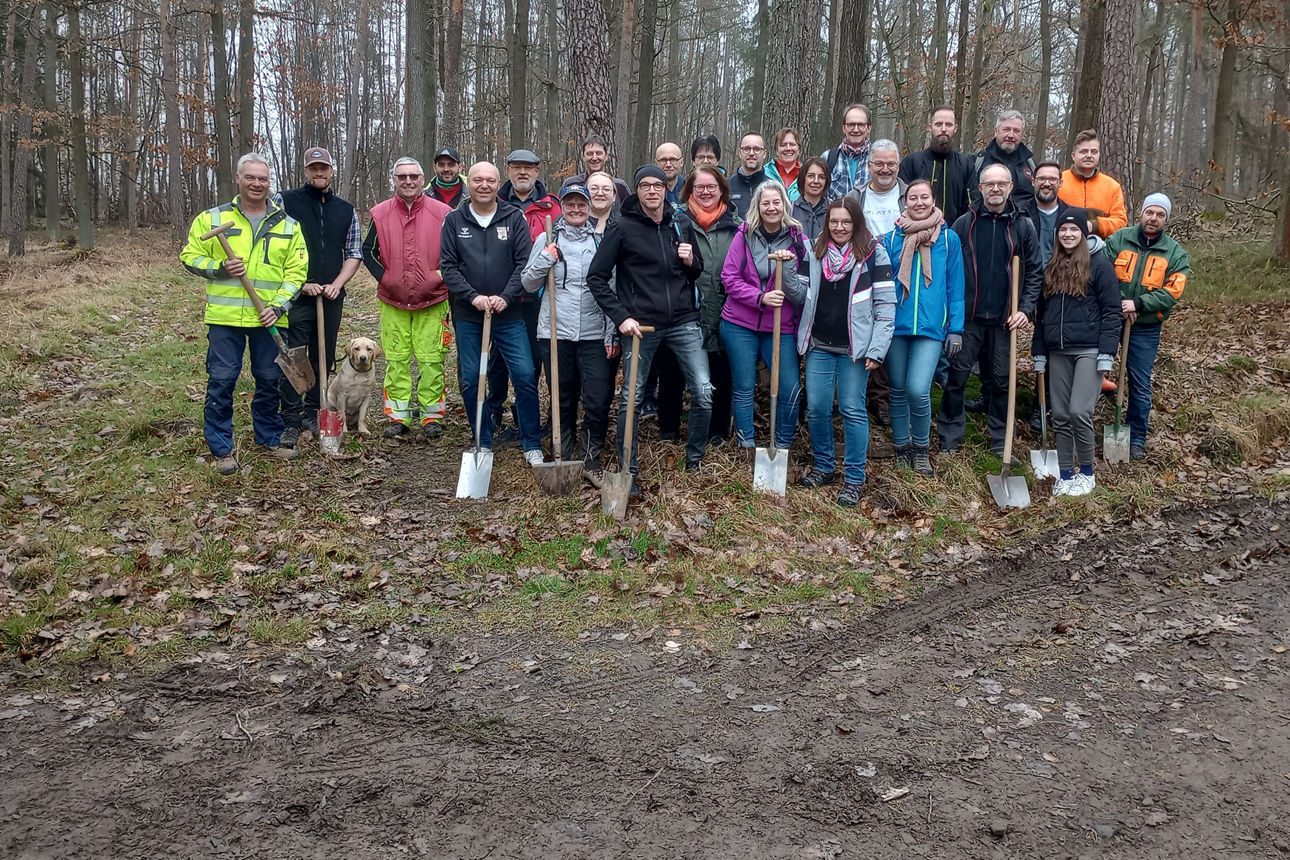 Ein Gruppe von Menschen steht in einem Waldgebiet und hat Baumsetzlinge und Spaten in der Hand.