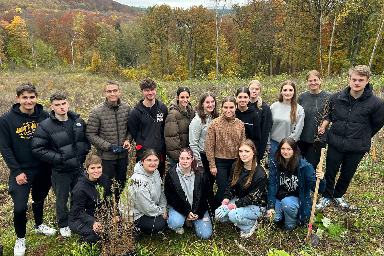 Auf einem Hügel vor einem Wald steht eine Gruppe von jungen Erwachsenen und Jugendlichen einige von ihnen haben Werkzeug 