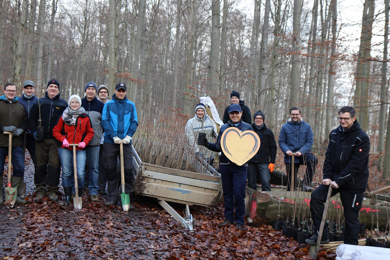 Einige Personen stehen in einem Wald vor einem Baumstumpf und halten Schaufeln in der Hand, einer halt ein Herz 