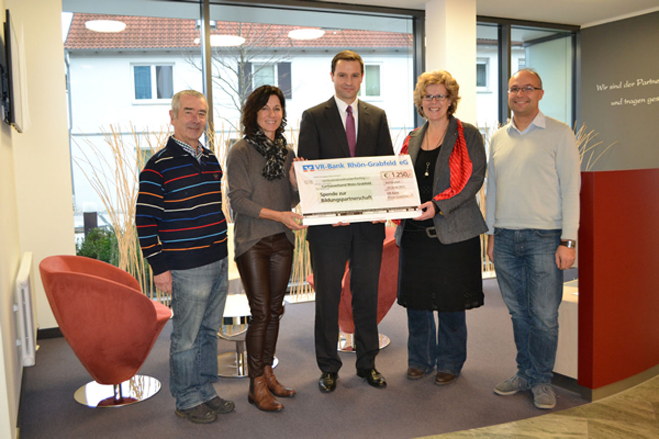 Vor einem Fenster stehen zwei Frauen und drei Männer, drei Personen in der Mitte halten einen Spendenscheck in der Hand 