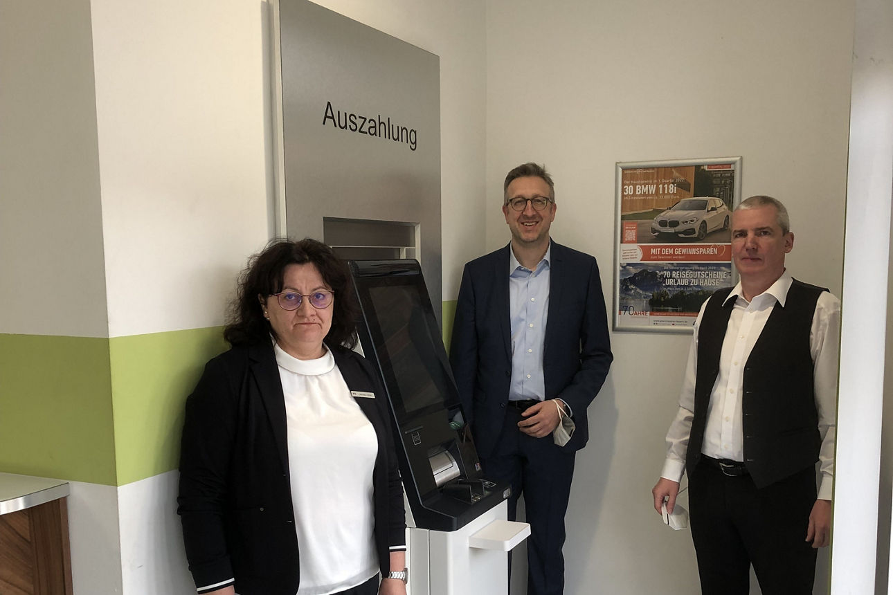 Drei Personen stehen in einem Raum neben einem Geldautomaten mit der Aufschrift „Auszahlung“