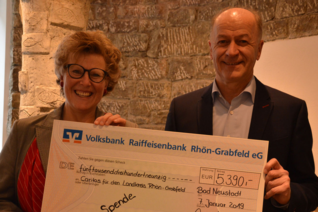 Vor einer Steinmauer steht ein Mann und eine Frau, die lächelnd einen Spendenscheck in der Hand halten 