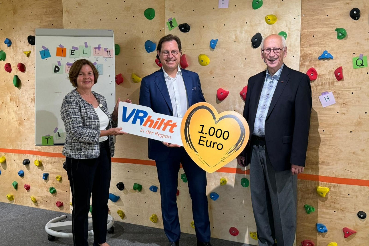Drei Personen stehen vor einer bunten Kletterwand, zwei von ihnen halten Schilder mit den Aufschriften VRhilft in der Region