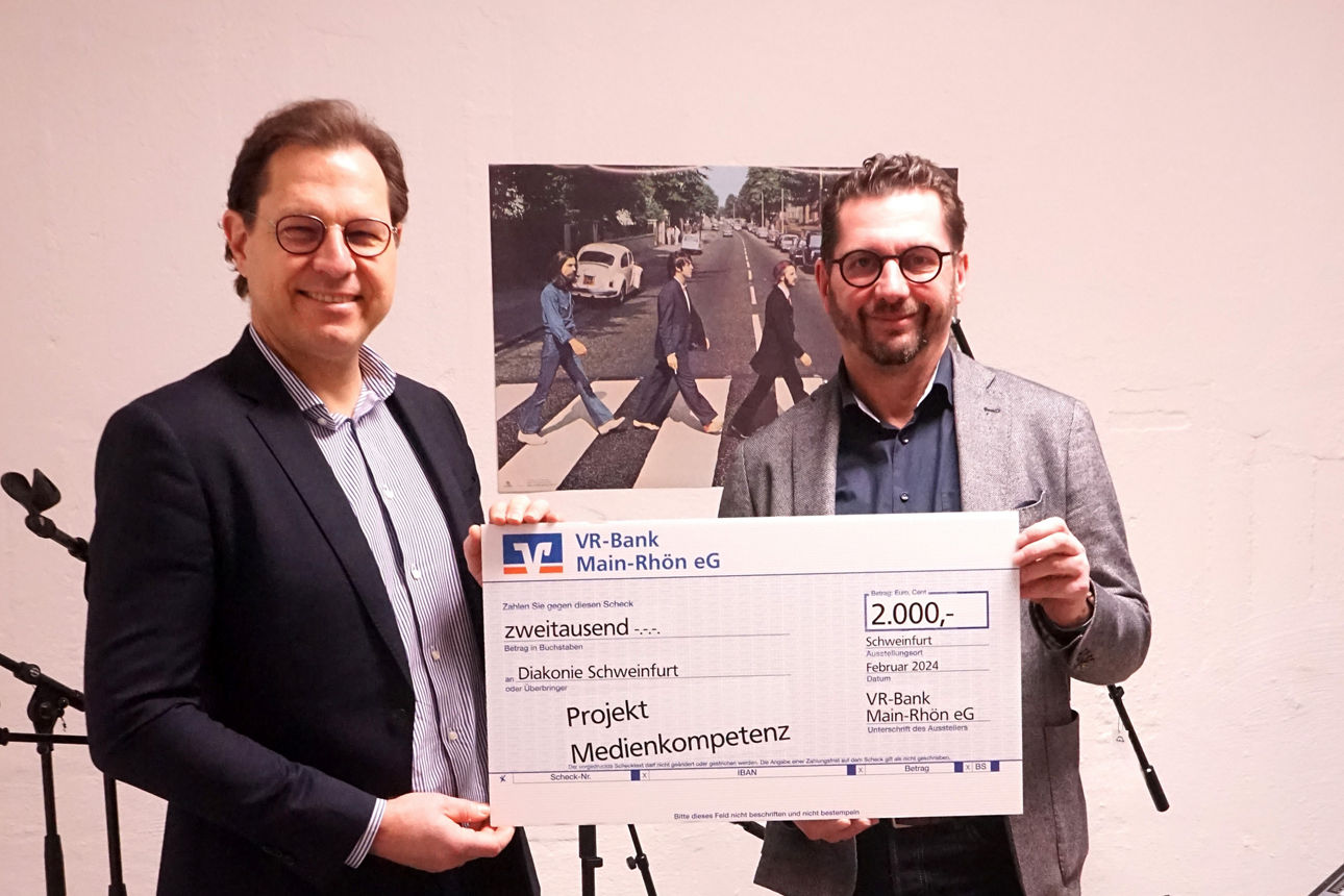 Zwei Männer stehen in einem Raum mit Mikrofonen und einem Mischpult und halten einen großen symbolischen Scheck