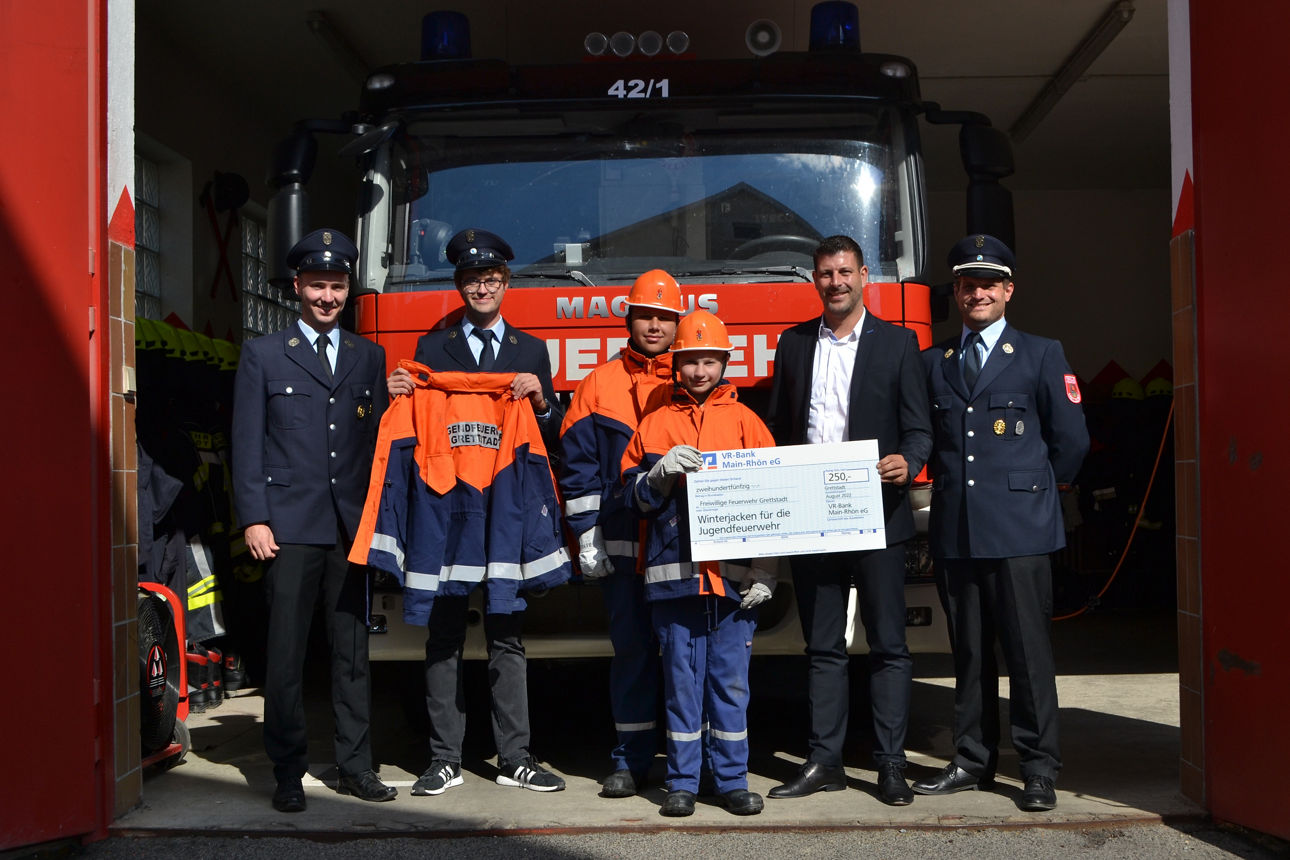 In dem Bild stehen sechs Personen vor einem großen roten Feuerwehrauto, zwei von ihnen tragen Jugendfeuerwehrkleidung